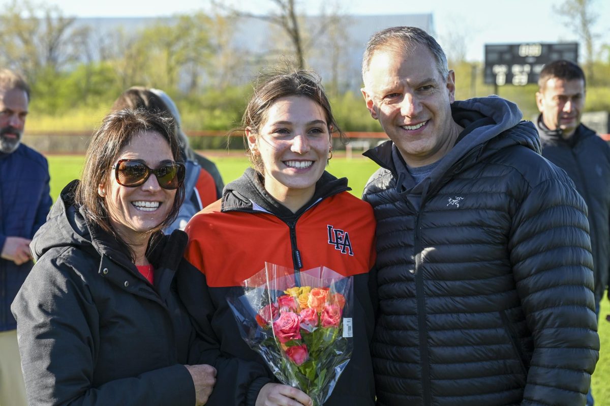 Kate Delia ‘25 smiles with her parents holding a bouquet of flowers on senior night.