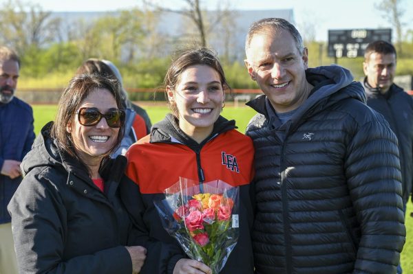 Kate Delia ‘25 smiles with her parents holding a bouquet of flowers on senior night.